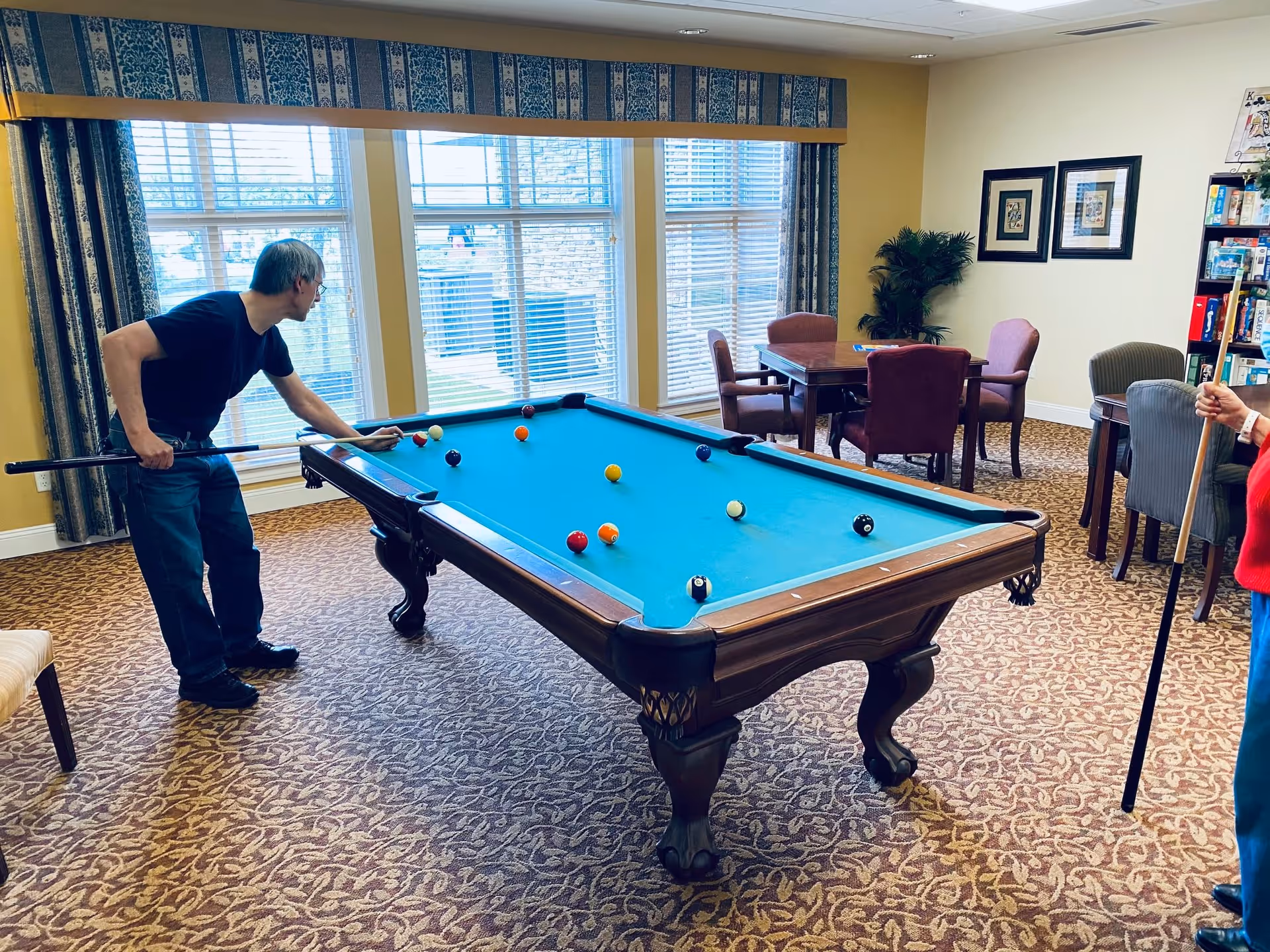 Two people playing pool in a well-lit game room with large windows, patterned carpet, and several tables and chairs in the background.