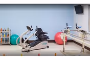 Physical therapy or exercise room with a recumbent stationary bike, parallel bars for walking support, colorful exercise balls, and a rack of small dumbbells against a light blue wall.
