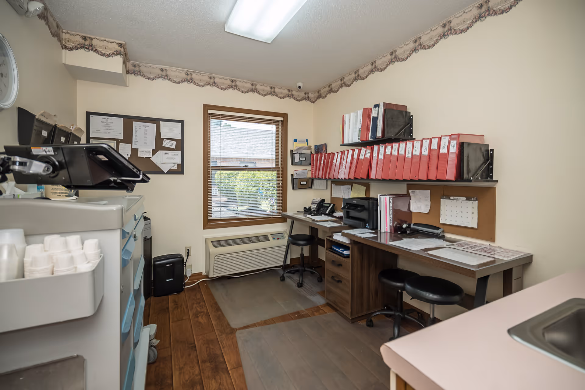 Small office room with wooden flooring and beige walls. There is a window with blinds letting in natural light. Two desks with black swivel stools are against the walls, holding office supplies, telephones, a printer, and numerous red binders neatly arranged on a wall-mounted shelf. A bulletin board with pinned papers is above one desk. A filing cabinet with trays and a small sink are also visible.