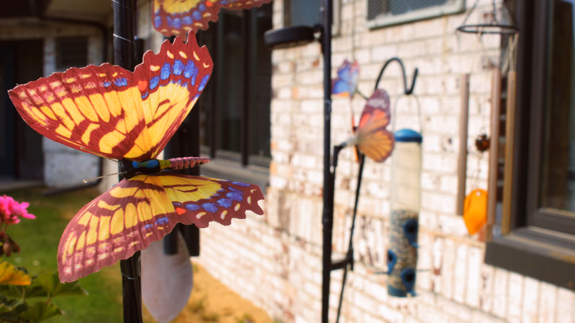 Close-up of colorful decorative butterfly garden stakes placed in front of a brick building wall with windows and a bird feeder visible in the background.