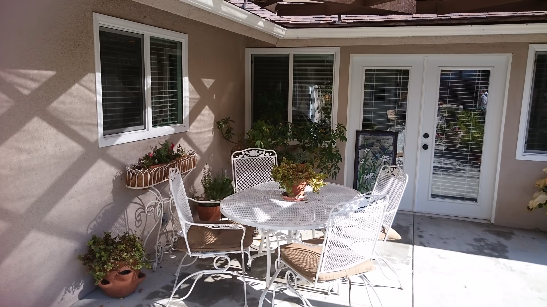 Outdoor patio area with a round white metal table and four matching chairs with beige cushions. Several potted plants are placed around the patio, including on the table and on the ground. The patio is adjacent to a beige building with white-framed windows and a white-framed glass door with blinds.