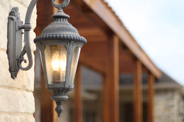 A close-up view of a vintage-style outdoor wall lantern mounted on a stone wall, with a wooden porch structure and roof blurred in the background.