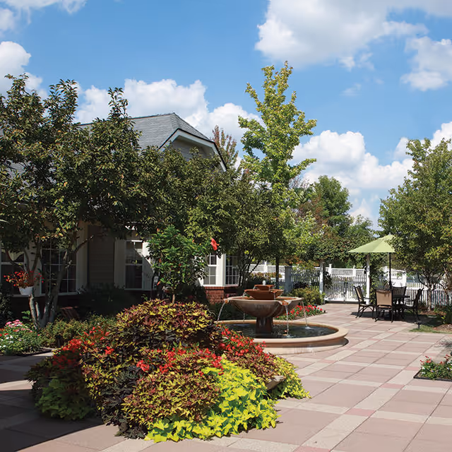 Outdoor courtyard area at Parc Provence featuring a central water fountain surrounded by lush greenery and colorful plants. There are several trees providing shade, a paved walkway, and a seating area with a table and chairs under a green umbrella. The sky is blue with scattered clouds.