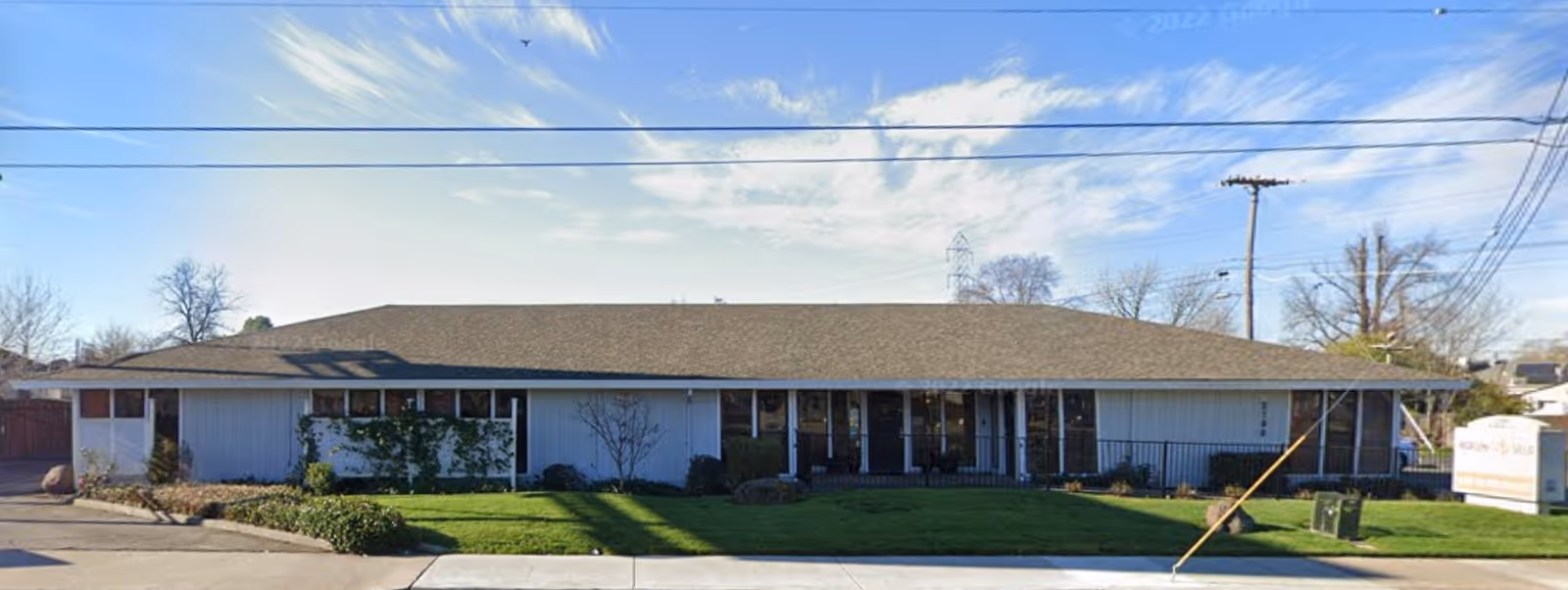 Single-story building with a brown shingle roof and light-colored exterior walls, surrounded by a well-maintained lawn and some shrubs. There are power lines overhead and a sign on the right side of the image indicating the facility name Marconi Villa.