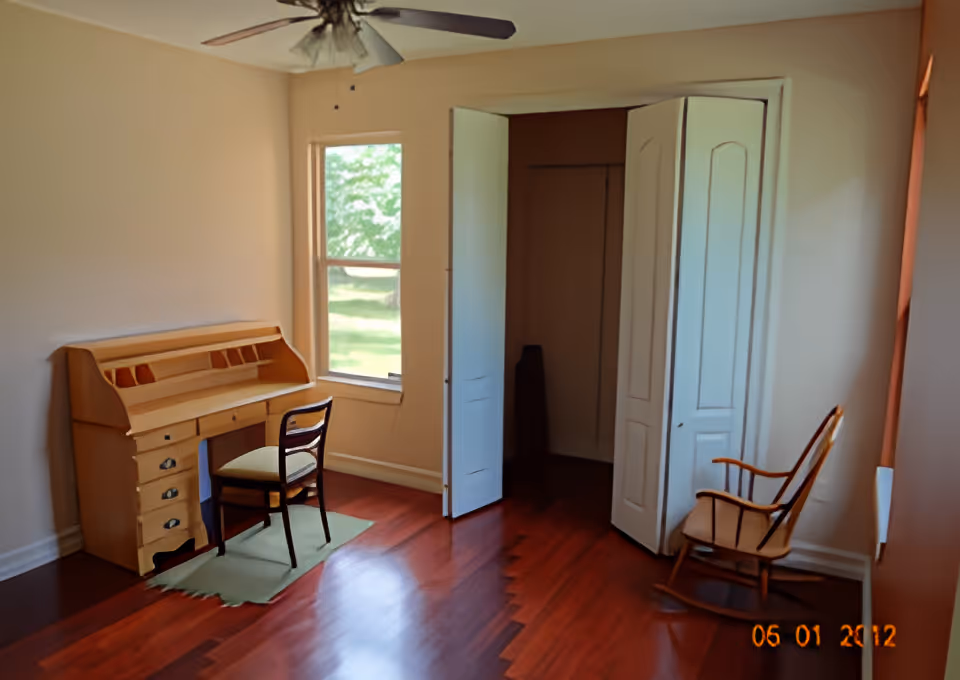 Empty bedroom with a wooden roll-top desk and chair by a window, a rocking chair, open closet doors, and a ceiling fan.