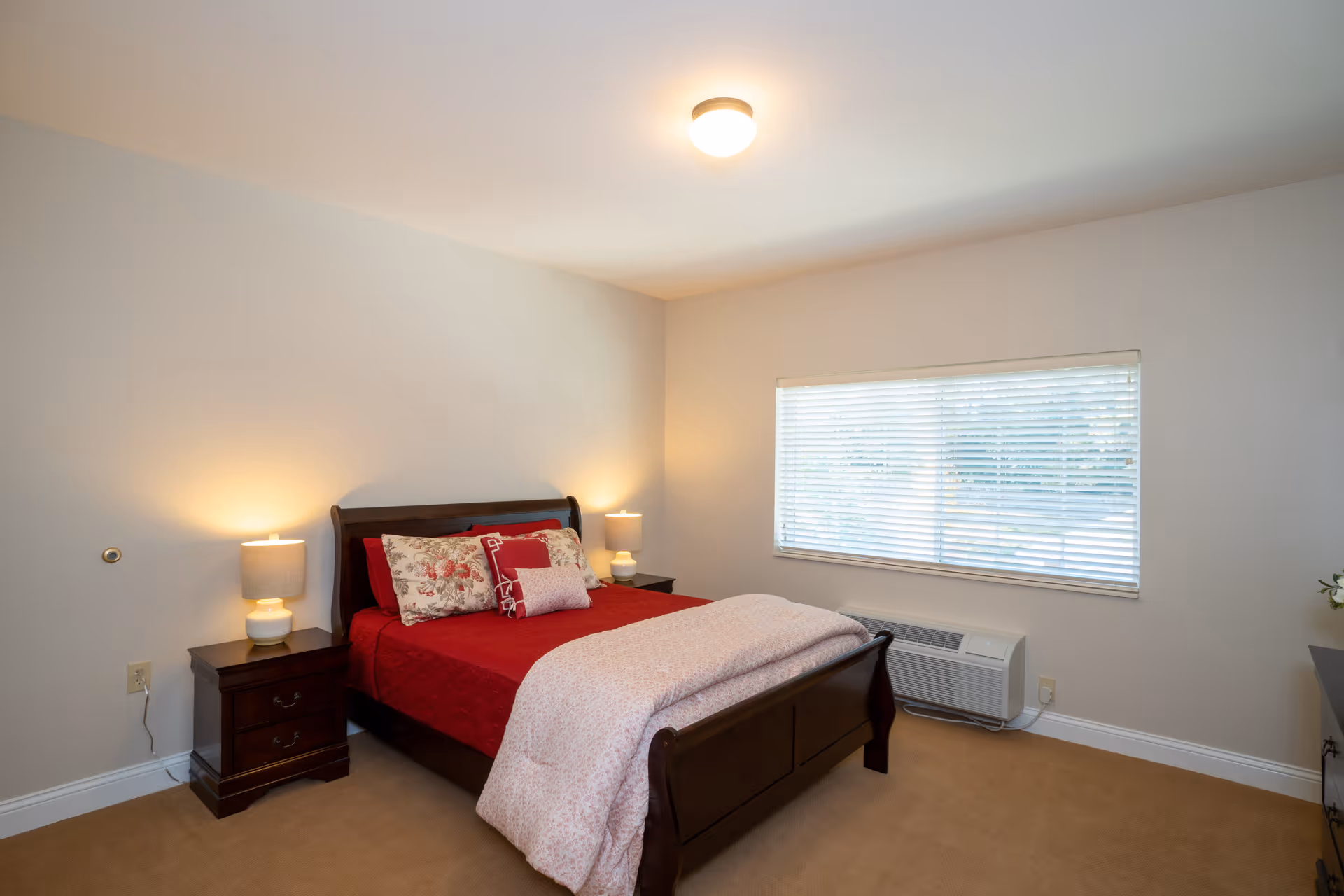 A bedroom with a wooden bed frame featuring red and floral bedding, two matching nightstands with lamps, a window with blinds, and a beige carpeted floor.