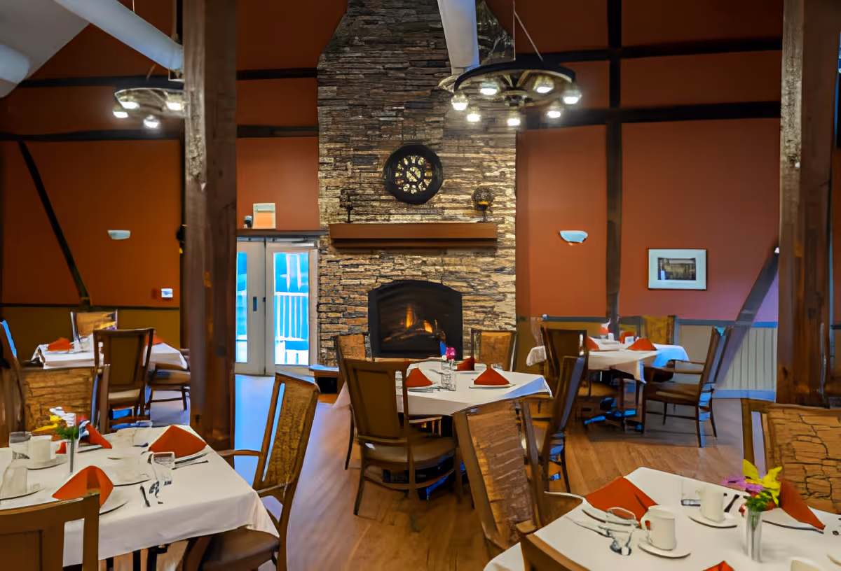 A cozy dining room with several tables set with white tablecloths, red napkins, cups, and glasses. The room features a large stone fireplace with a fire burning, wooden beams, and warm orange walls. There are chairs around the tables and a chandelier hanging from the ceiling.