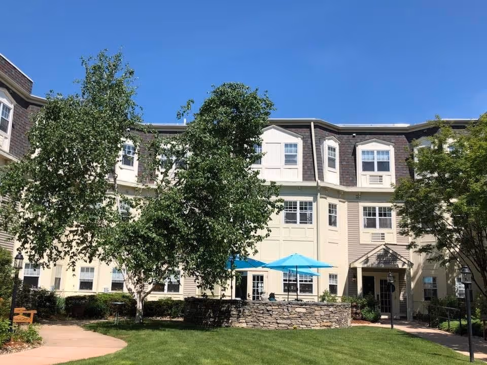 Exterior view of a multi-story senior living facility building with beige siding and brown shingles under a clear blue sky. In front of the building is a green lawn with a stone patio area featuring blue umbrellas and outdoor seating. Trees and shrubs surround the area, and a paved walkway leads to the entrance.