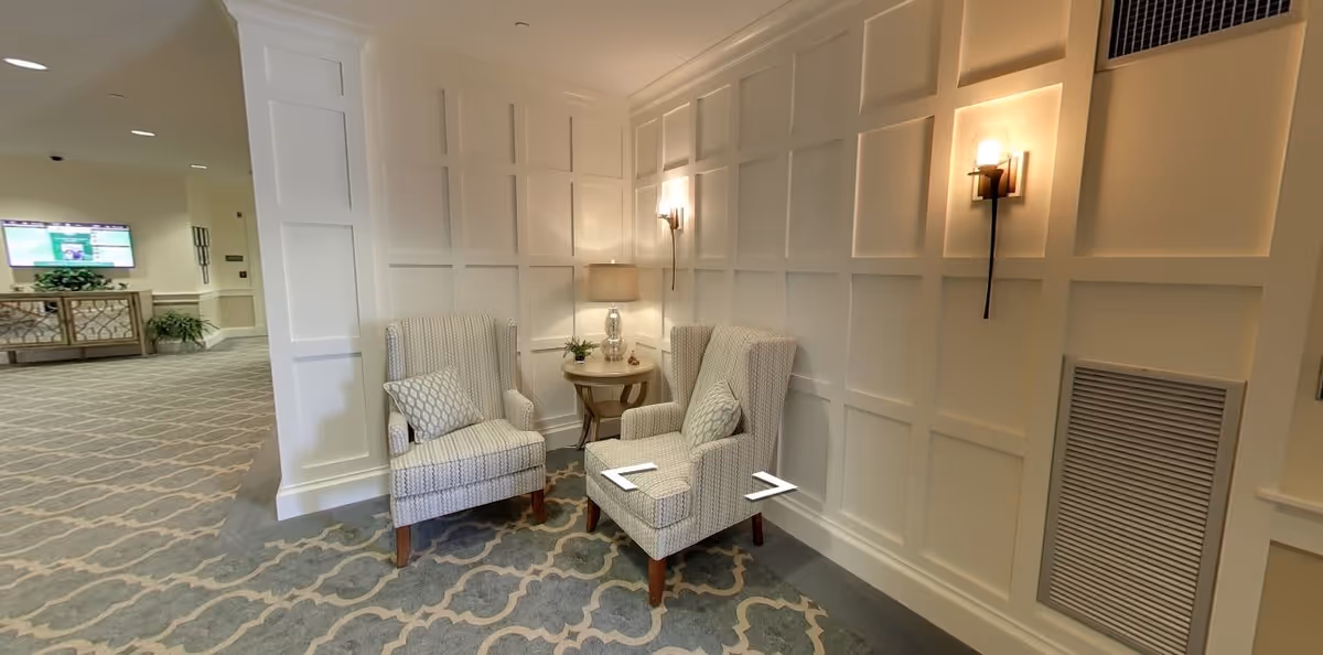 A cozy seating area in a hallway with two patterned armchairs, each with a decorative pillow, positioned around a small round wooden table with a lamp and a small plant. The walls have white paneling and two wall sconces provide warm lighting. The floor is carpeted with a patterned design, and in the background, there is a TV mounted on the wall and a console table with plants.
