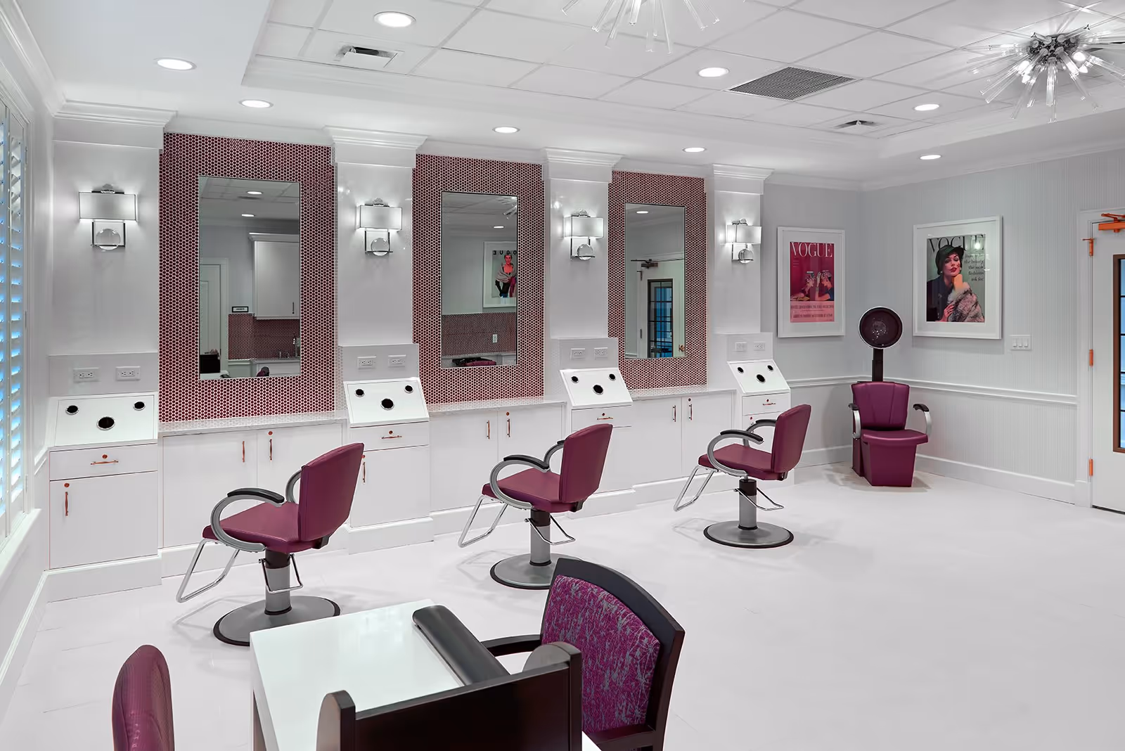 A modern salon area with three purple salon chairs in front of large mirrors mounted on a white wall with purple mosaic tile accents. The room has white walls, bright lighting, and two framed Vogue magazine covers on the right wall. There is also a purple salon dryer chair in the corner and a white table with a purple chair in the foreground.