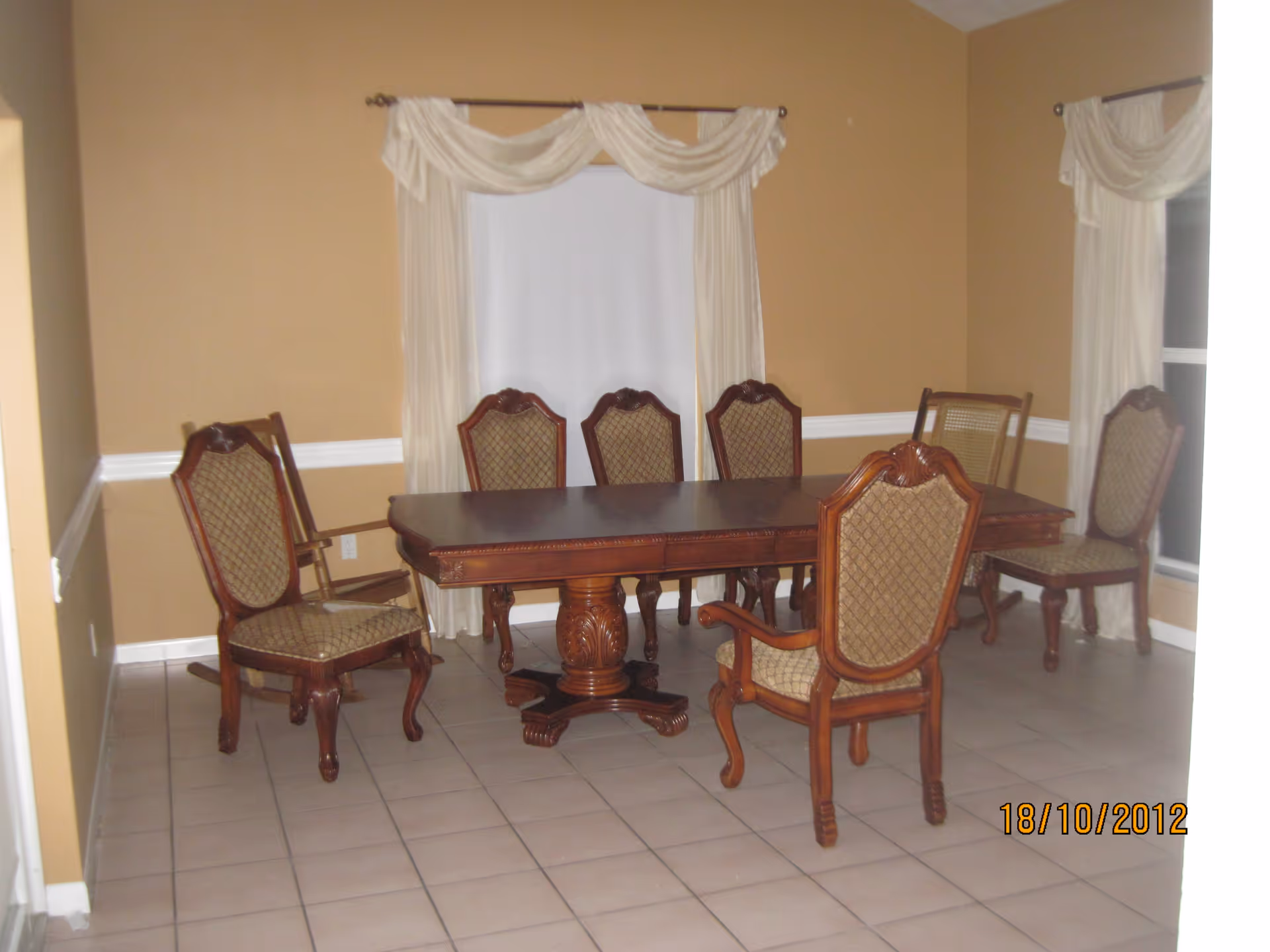 A dining room with a rectangular wooden dining table surrounded by six upholstered chairs with wooden frames. The room has beige walls, white tile flooring, and two windows with cream-colored curtains.