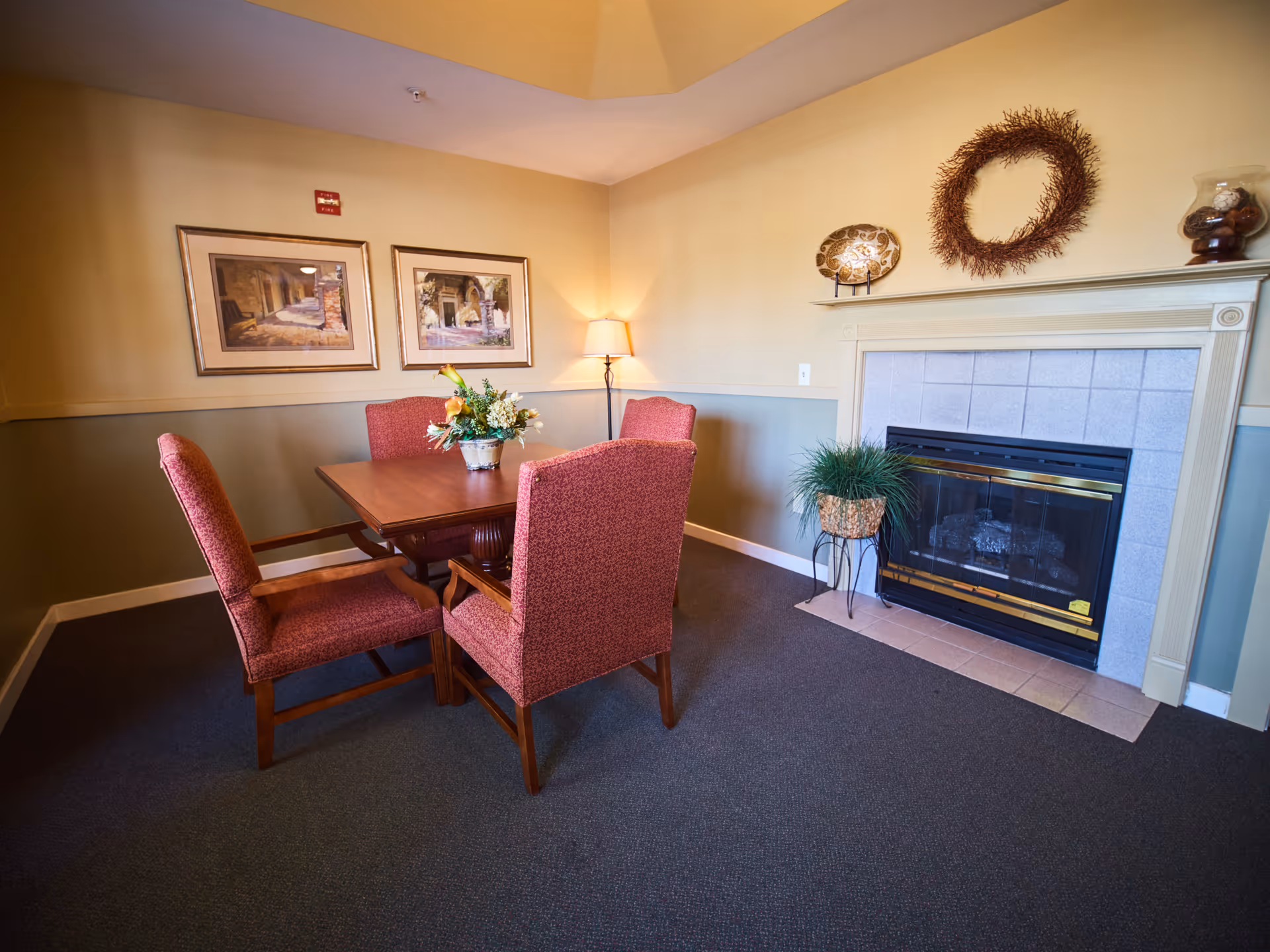 A cozy interior room with a wooden table surrounded by four red upholstered chairs. The room features a fireplace with a decorative wreath above it, a potted plant on a stand next to the fireplace, two framed pictures on the wall, and a floor lamp in the corner providing warm lighting.