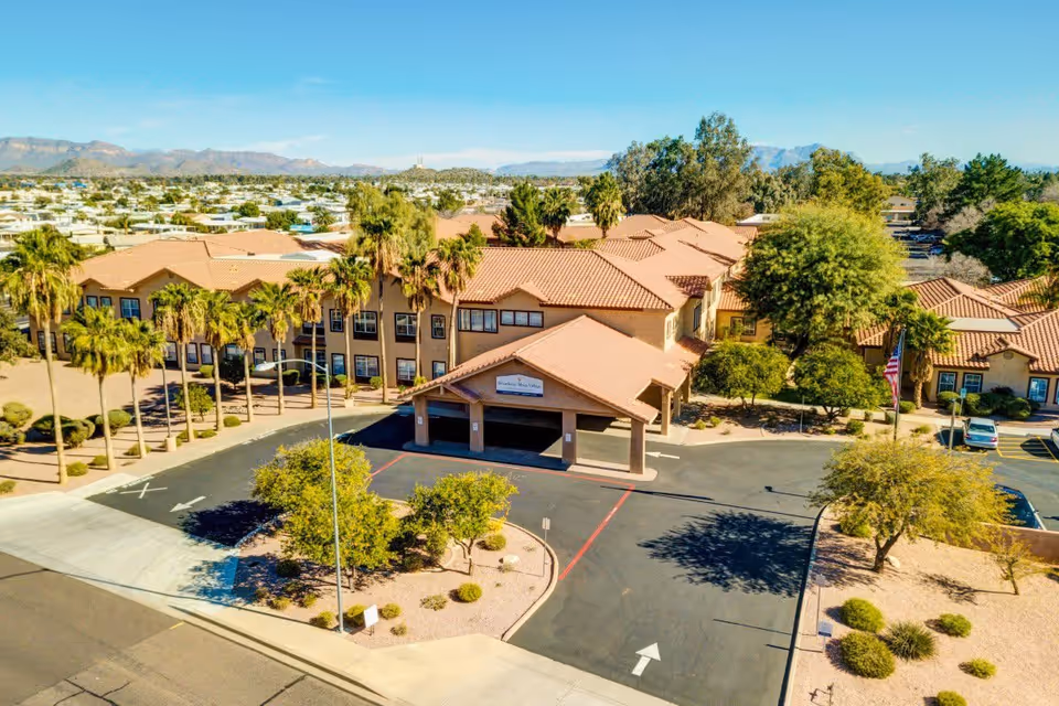 Aerial view of Pegasus Landing of Mesa, a senior living facility with a large building featuring a terracotta tiled roof, surrounded by palm trees and desert landscaping. The driveway leads to a covered entrance, and mountains are visible in the background under a clear blue sky.