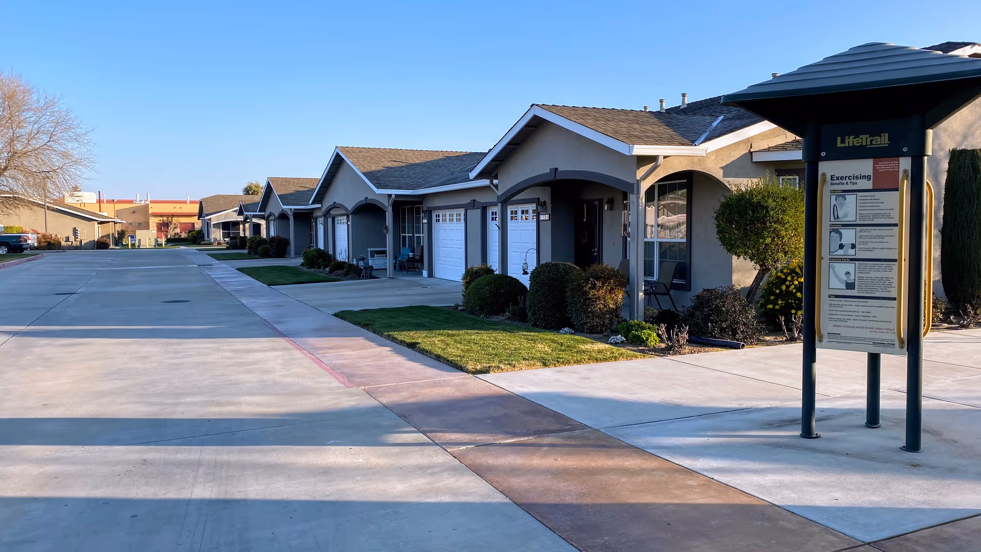 A row of single-story residential buildings with garages and small front lawns along a wide concrete driveway. There is a LifeTrail outdoor exercise station with instructions visible on the right side of the image. The sky is clear and the scene is well-lit by sunlight.