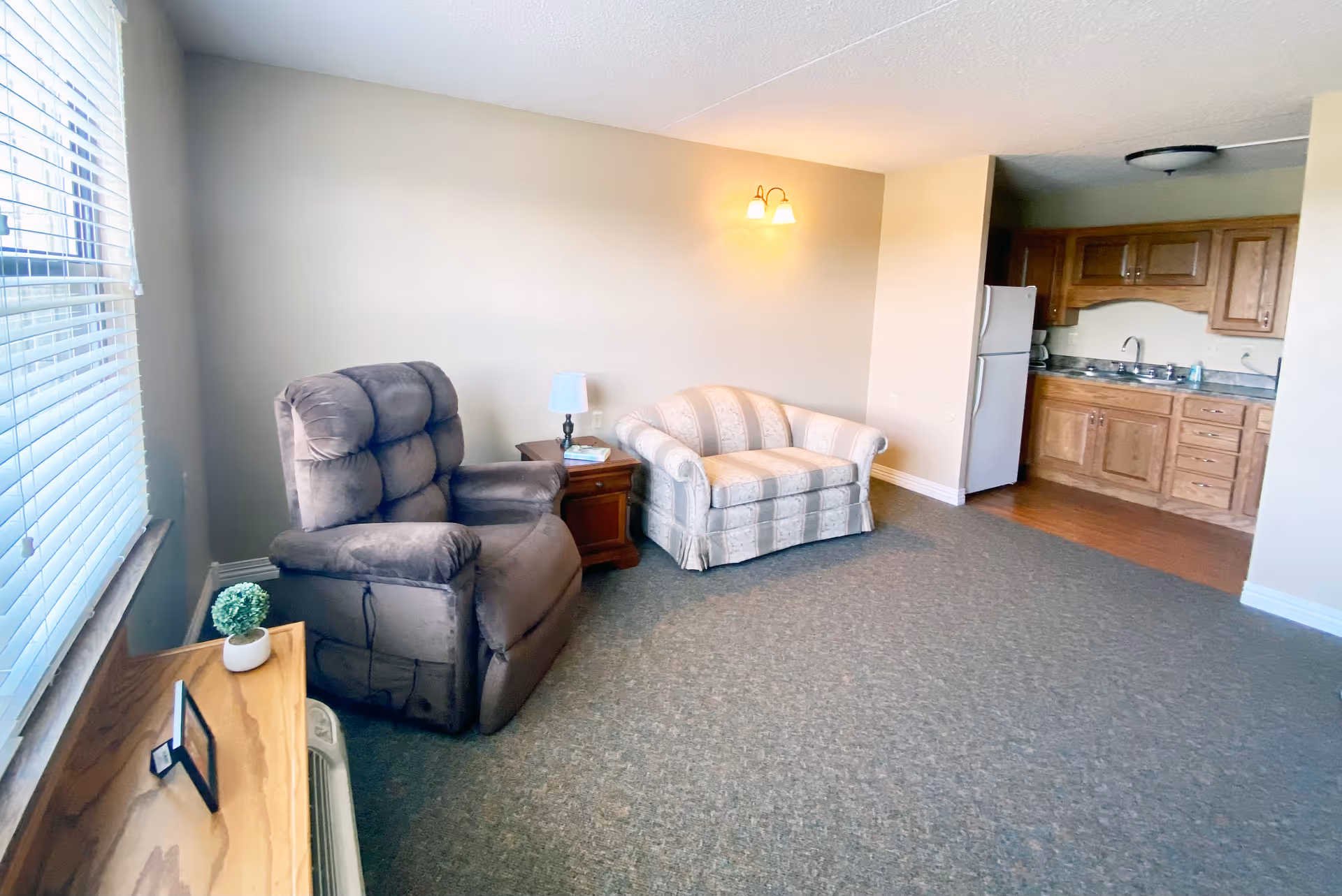 A small living area with a gray recliner chair and a striped loveseat sofa. There is a wooden side table with a lamp and books between the chairs. To the right, there is a kitchenette with wooden cabinets, a sink, and a white refrigerator. A window with blinds is on the left side, letting in natural light.