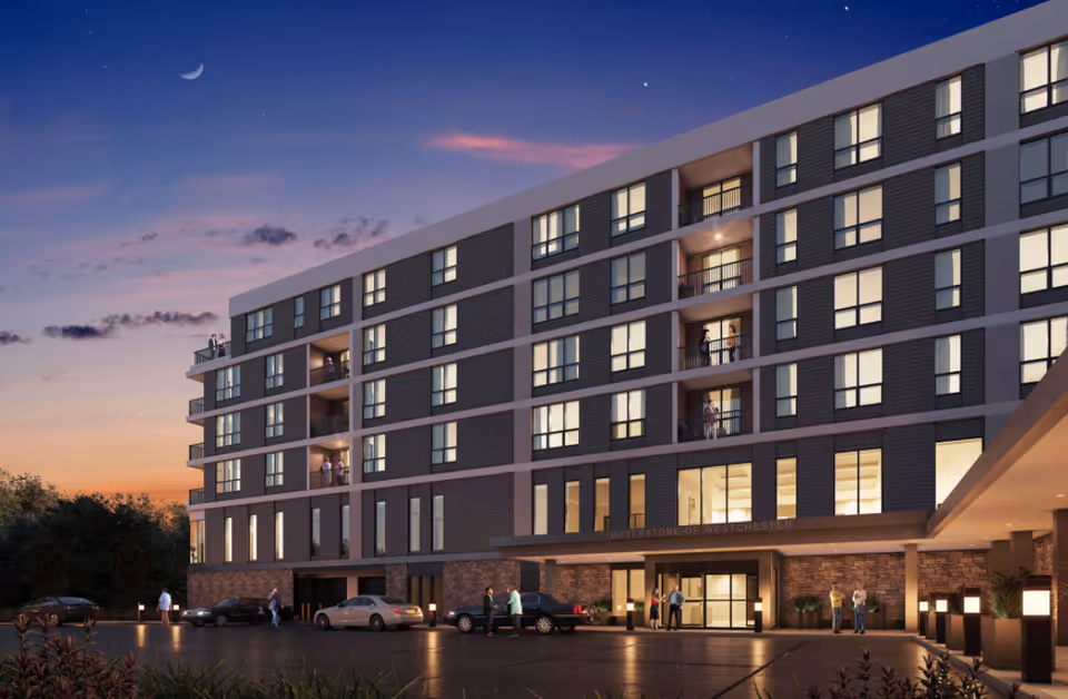 Exterior view of a modern multi-story senior living facility named Waterstone of Westchester at dusk, with several people standing on balconies and near the entrance, cars parked in front, and a crescent moon visible in the sky.