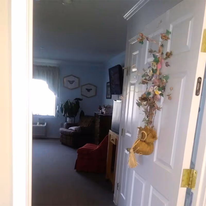 View into a living room area from an open doorway. The door has a decorative hanging with dried flowers and a small burlap sack. Inside the room, there is a window with curtains, a potted plant, a plaid armchair, a red chair, a wooden cabinet, and a wall-mounted TV. Two framed pictures hang on the wall.