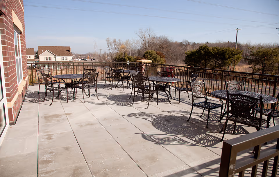 Outdoor patio area with several black metal tables and chairs arranged on a tiled floor, surrounded by a metal railing. In the background, there are trees and a few houses under a clear sky.