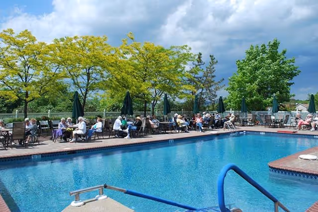 Outdoor swimming pool with chairs and tables along the deck where many residents sit under umbrellas, with trees and a cloudy sky in the background.