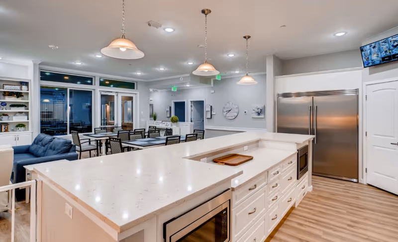 A modern and spacious interior area featuring a large white kitchen island with drawers and built-in appliances, pendant lights hanging from the ceiling, a stainless steel refrigerator, and a dining area with tables and chairs. There is a blue couch near the dining area and large windows and doors in the background.