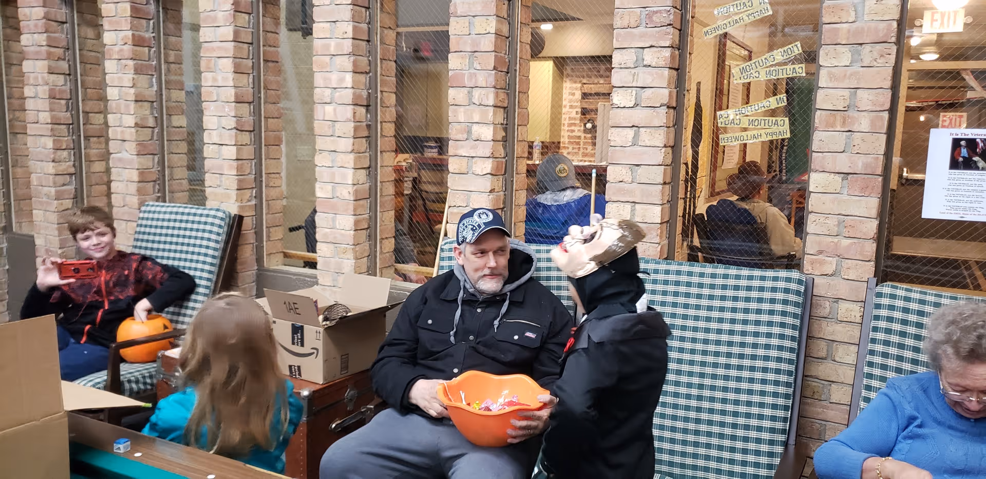 A group of people, including children and an older man, sitting in a room with brick walls and large windows. The older man is holding an orange bowl filled with candy, and one child is wearing a Halloween mask. There are boxes and pumpkins around, suggesting a Halloween-themed gathering.