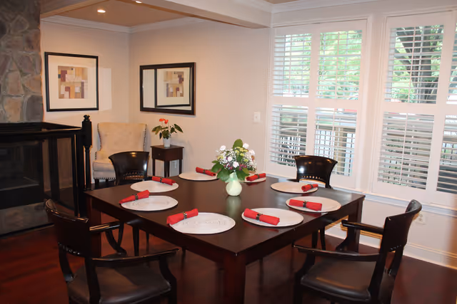 A dining room with a dark wooden table set for six people with round placemats and red napkins. There are six dark wooden chairs around the table. A vase with flowers is placed in the center of the table. The room has large windows with white blinds, a stone fireplace on the left, a beige armchair, a small side table with a plant, and two framed abstract artworks on the wall.