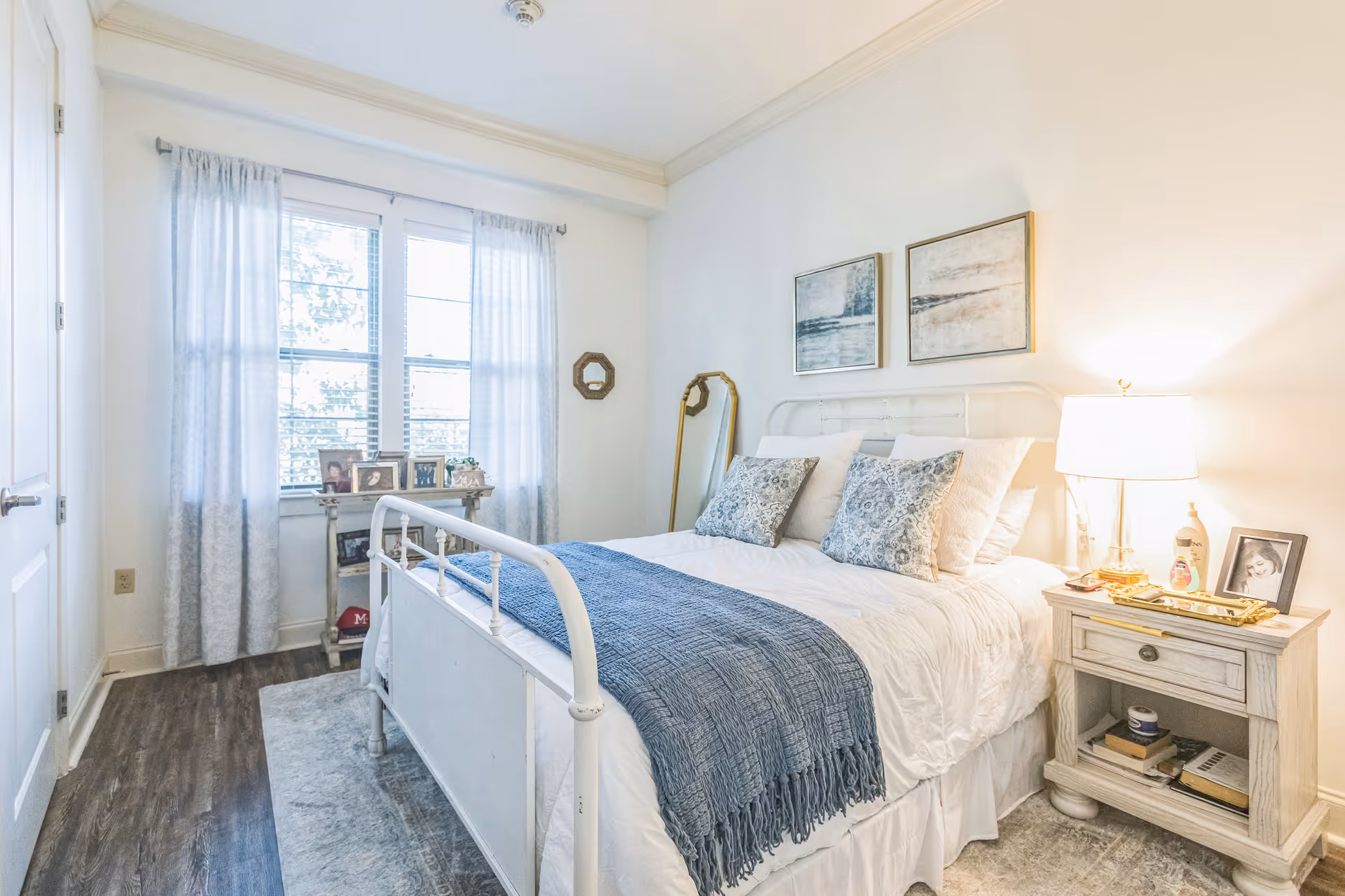 A bright and cozy bedroom with a white metal bed frame, white bedding, and a blue throw blanket. There are two patterned pillows on the bed. A wooden nightstand with a lamp, framed photo, and some items is on the right side of the bed. Two abstract paintings hang on the wall above the bed. A window with sheer curtains lets in natural light, and a small table with framed photos is placed beneath the window. The floor is wooden with a light-colored rug beside the bed.