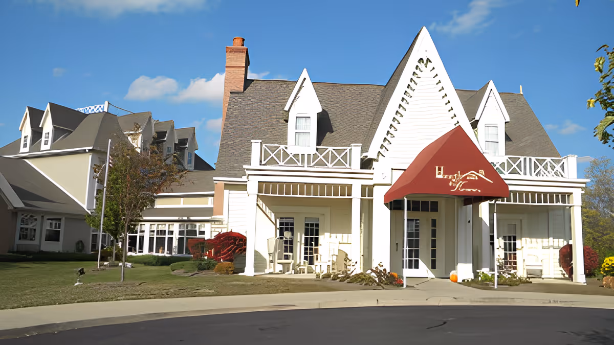 Front exterior view of a senior assisted living facility building with a steep roof, white siding, and a red awning over the entrance. There are small trees and shrubs in the landscaped lawn area in front of the building under a clear blue sky.