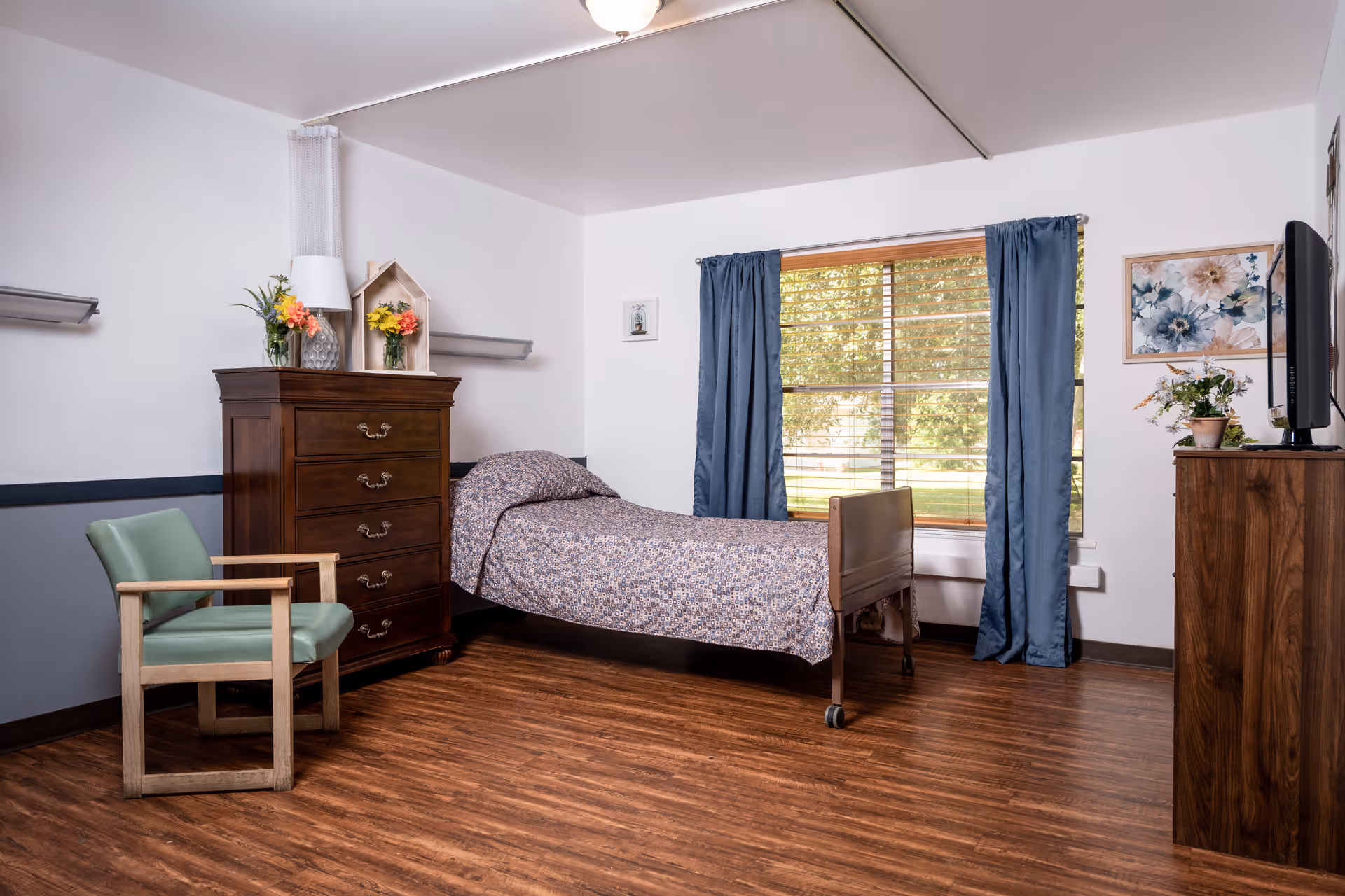 A senior living facility bedroom with a single bed covered in a patterned bedspread, a wooden dresser with flowers and decorative items on top, a green cushioned chair, a window with blue curtains showing a view of trees outside, and a wooden TV stand with a television and a potted plant.