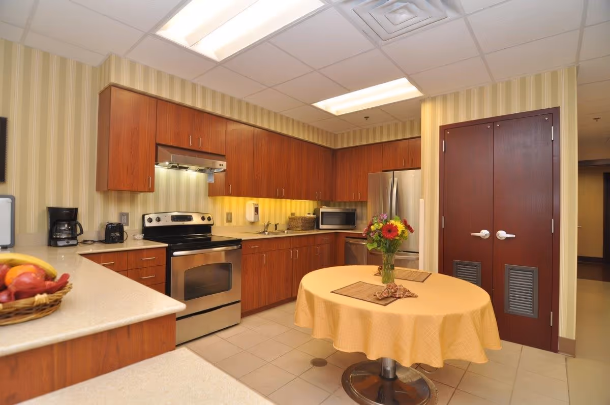 A clean kitchen with wooden cabinets, stainless steel appliances including an oven, microwave, and refrigerator. There is a round table in the center covered with a yellow tablecloth and a vase of colorful flowers on top. A basket of fruit is visible on the counter to the left. The walls have vertical striped wallpaper and there is a double door with dark wood finish on the right side.