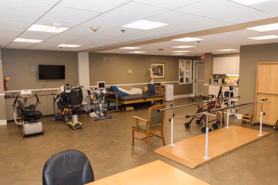 A rehabilitation room with exercise equipment including stationary bikes, a therapy bed, parallel bars for walking practice, and walkers. The room has beige walls, a mounted TV, a chair, and a small kitchenette area in the background.