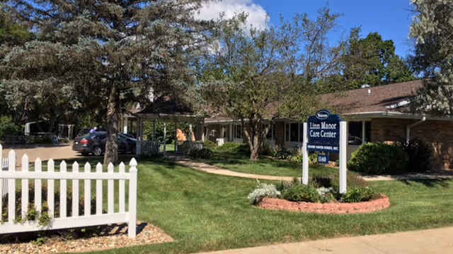 Exterior view of Linn Manor Care Center showing a single-story building with a brown roof surrounded by trees and greenery. A white picket fence is visible on the left side, and a blue sign with the facility's name stands in a landscaped area with grass and shrubs. A car is parked near the building under a covered area.