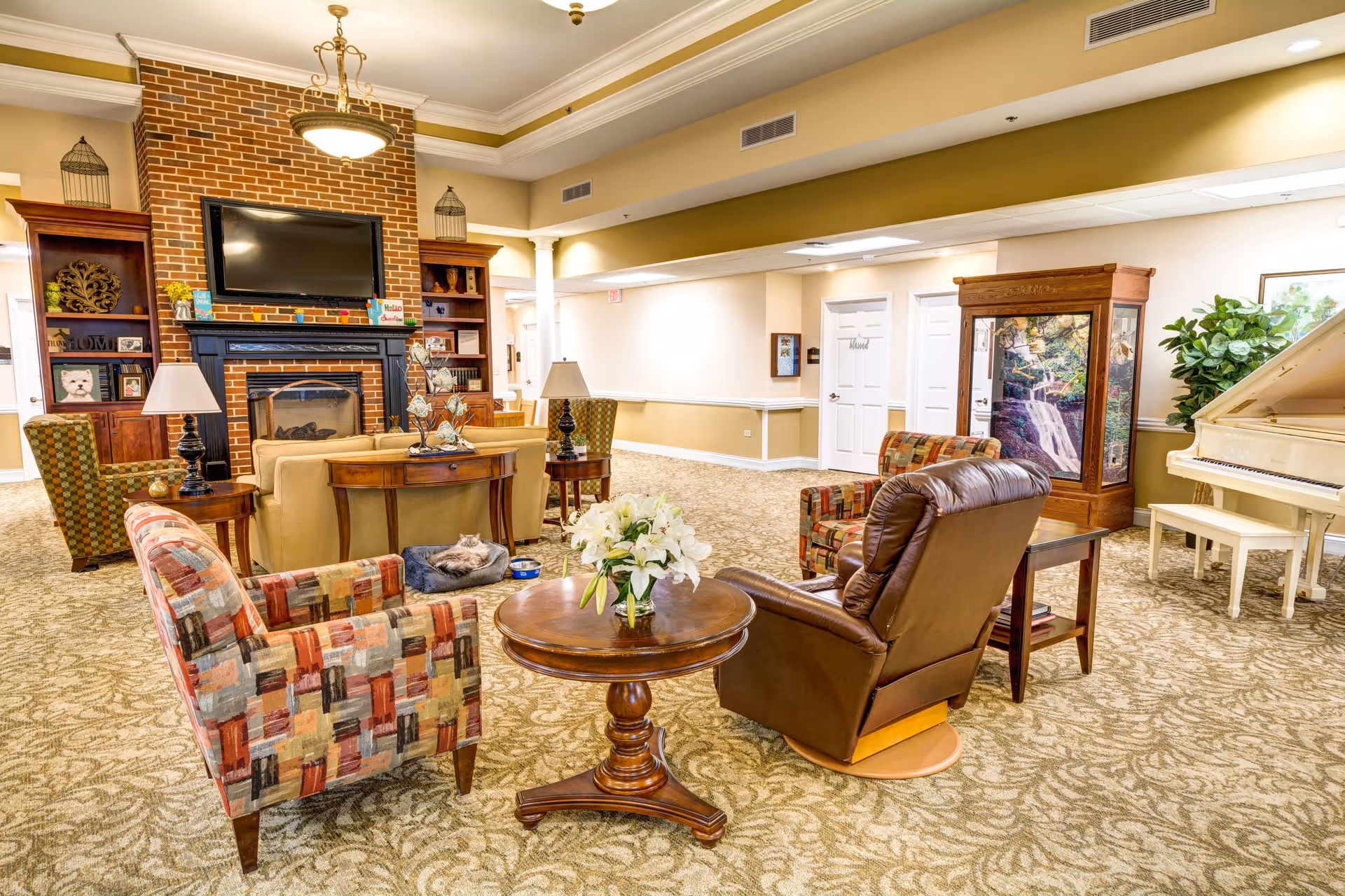 A cozy senior living facility common area with patterned armchairs, a leather recliner, a round wooden table with a flower vase, a beige sofa, and a brick fireplace with a mounted flat-screen TV. There are built-in wooden shelves on either side of the fireplace, a white piano on the right, and a decorative cabinet with a waterfall image. The room has warm lighting and carpeted floors.