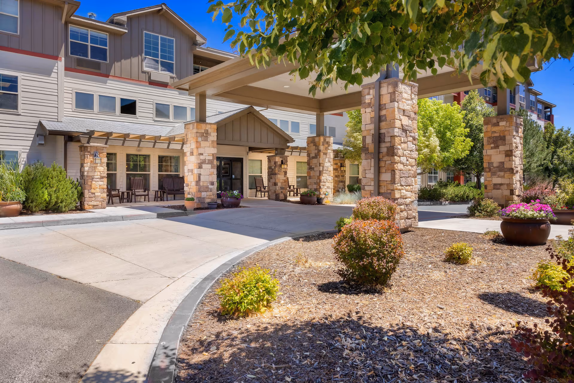 Entrance of MorningStar Senior Living of Sparks featuring a covered drop-off area supported by stone pillars, surrounded by landscaped bushes and potted flowers under a clear blue sky.