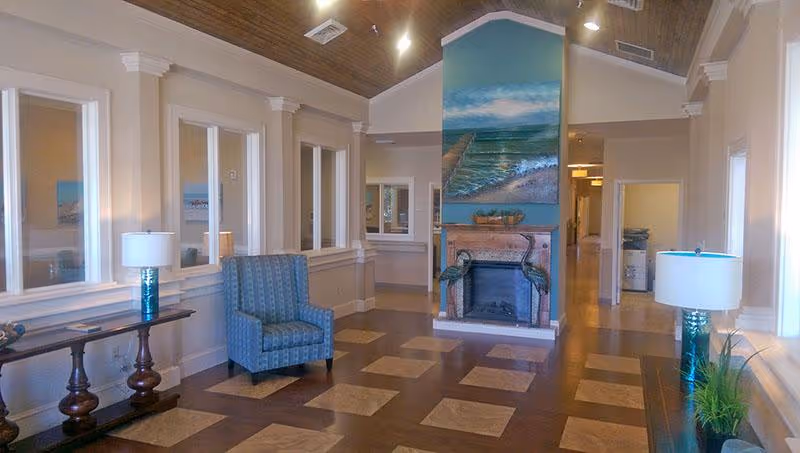 Bright communal lounge with a decorative fireplace, a blue armchair, side tables with lamps, and a vaulted wood ceiling.