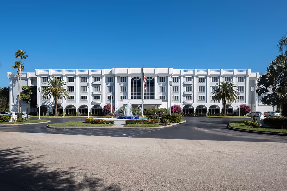 Front exterior view of a large white multi-story building with multiple windows and balconies, surrounded by palm trees and landscaping. A circular driveway with a fountain and an American flag is in front of the building under a clear blue sky.