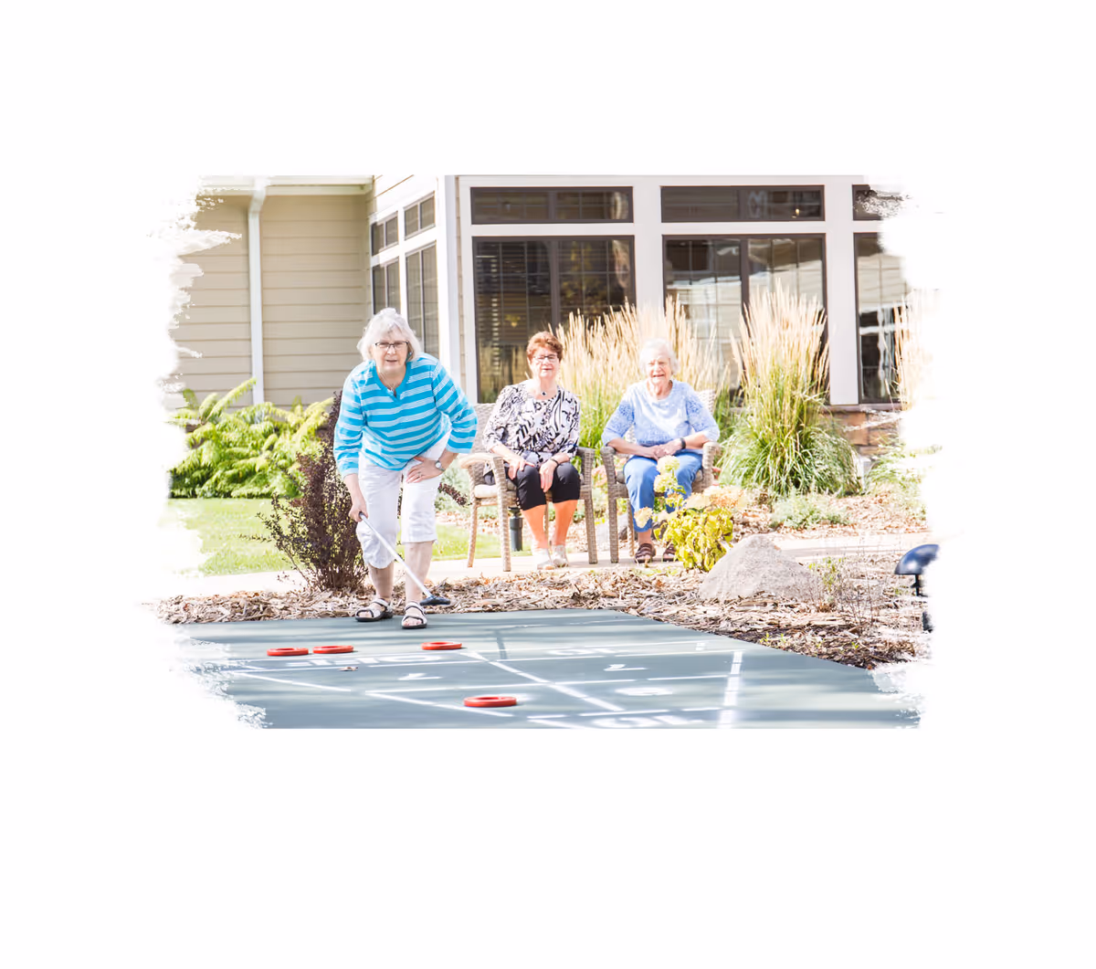 Three elderly women enjoying a game of shuffleboard outdoors at a senior living facility. One woman in a blue and white striped shirt is actively playing, while the other two women sit on chairs watching and smiling. The background shows a building with large windows and landscaping with plants and rocks.