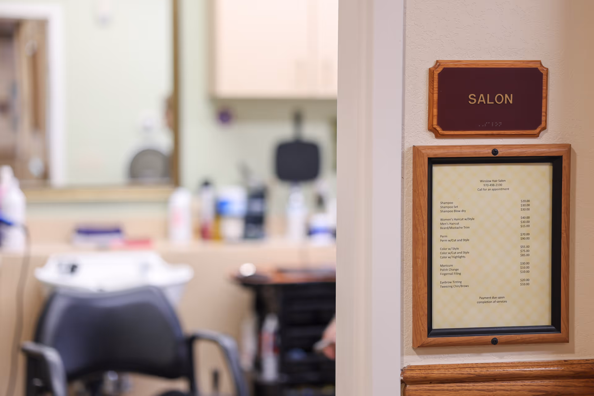 Interior view of a salon area in a senior living facility, showing a black salon chair and a wash basin in front of a mirror with various hair care products on the counter. On the wall outside the salon, there is a sign that reads 'SALON' and a framed price list for salon services.