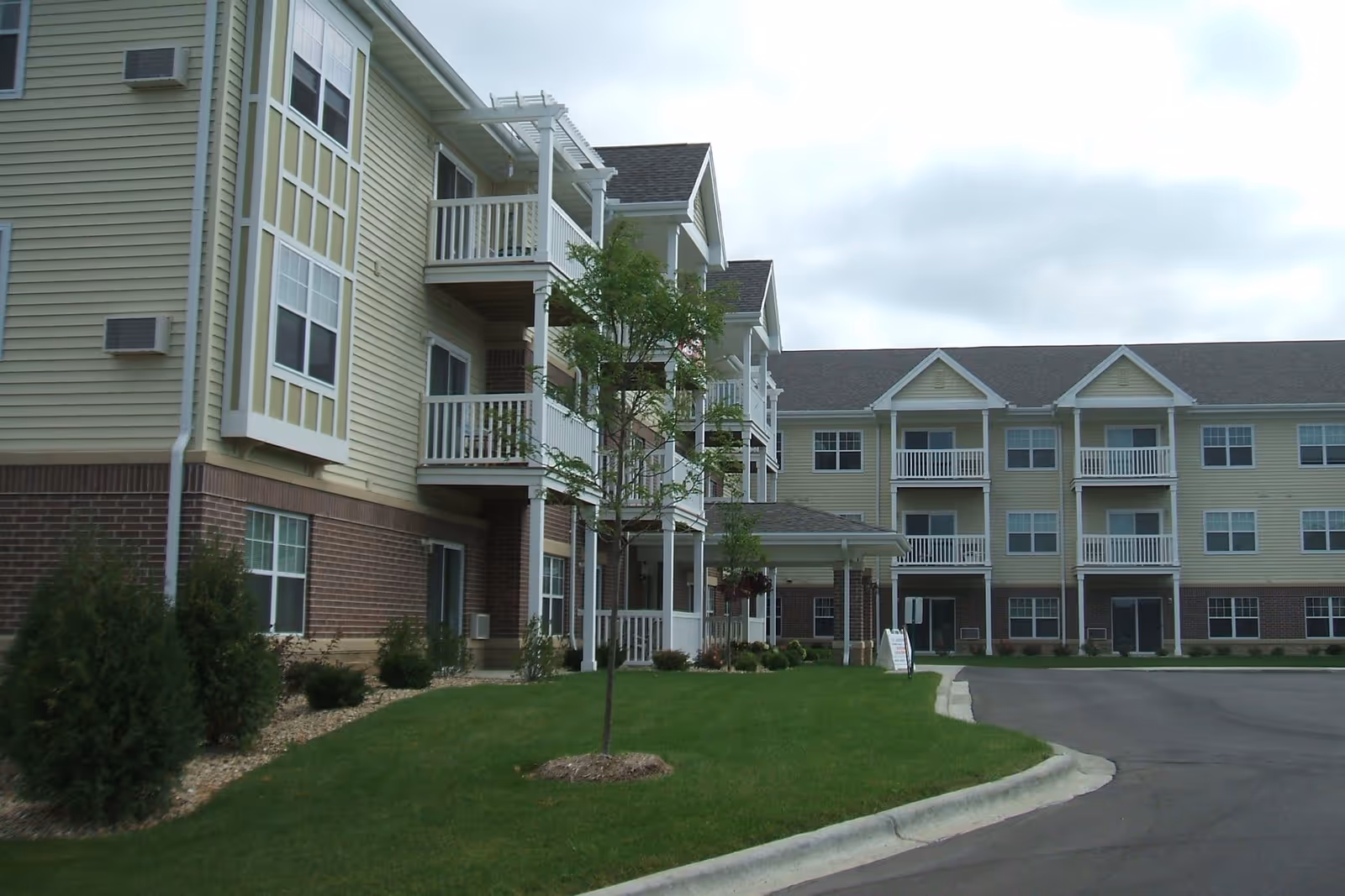 Exterior view of a three-story senior apartment building with beige siding and brick accents. The building features multiple balconies, a covered entrance, and a well-maintained lawn with small trees and shrubs. The sky is cloudy.
