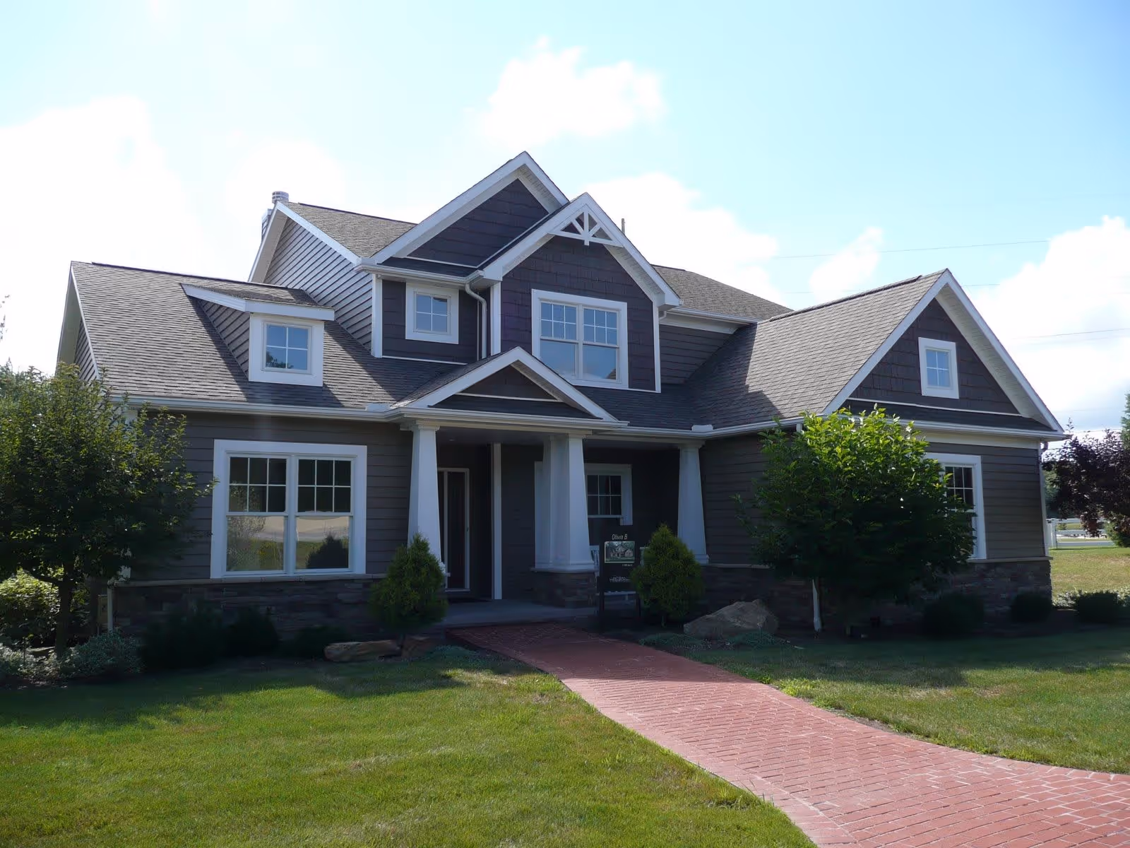 Front exterior view of a two-story residential building with gray siding, white trim, and a covered porch. There is a brick walkway leading up to the entrance, surrounded by green grass and small trees.