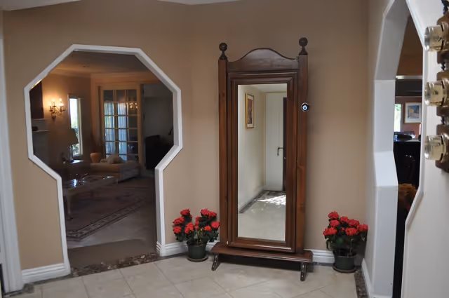 Foyer with a wooden full-length mirror flanked by potted red flowers and two octagonal openings showing a sitting room.