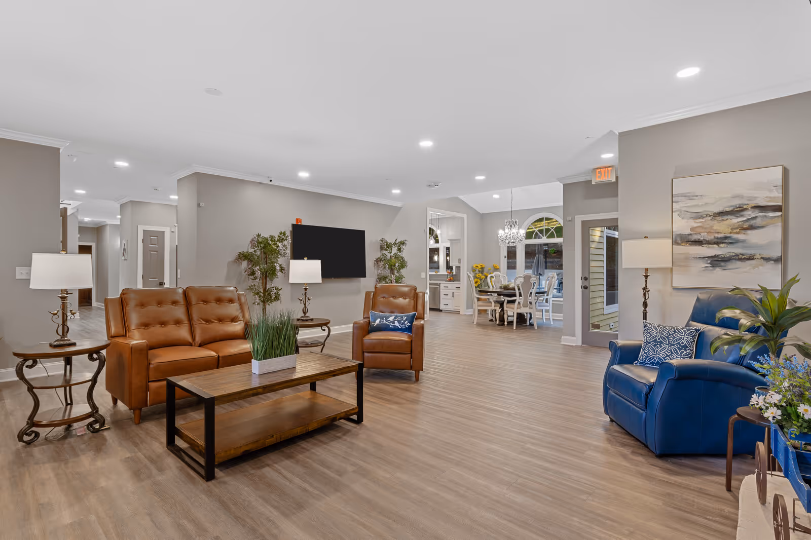 A spacious senior living common area with brown leather sofa and armchair, a blue armchair, wooden coffee table with a plant, side tables with lamps, a wall-mounted TV, and a dining area with a chandelier and white chairs in the background.
