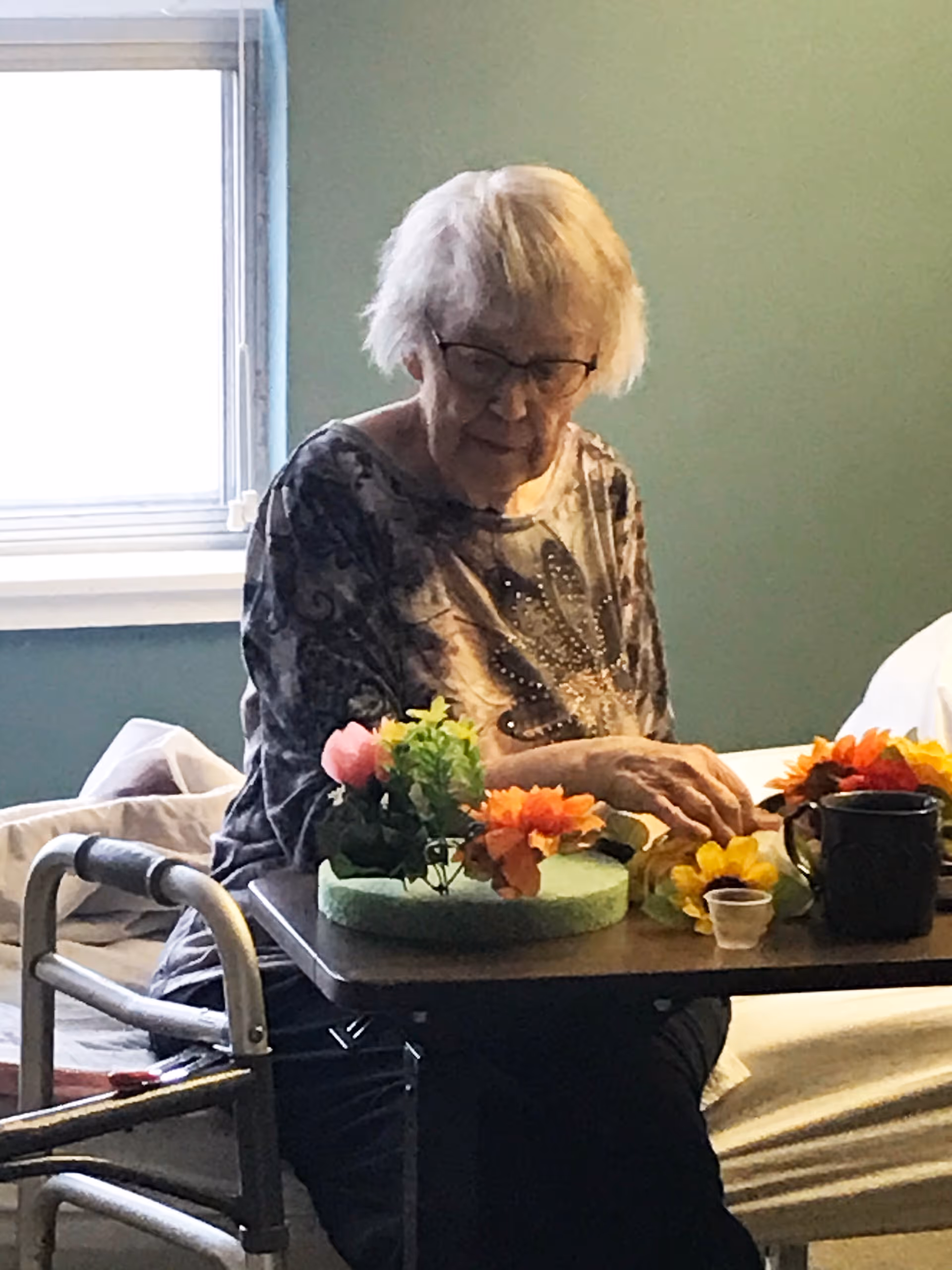 An elderly woman with glasses sitting at a table in a room with a window. She is arranging colorful artificial flowers on a green foam base. A walker is positioned next to her, and a bed with white bedding is visible in the background.