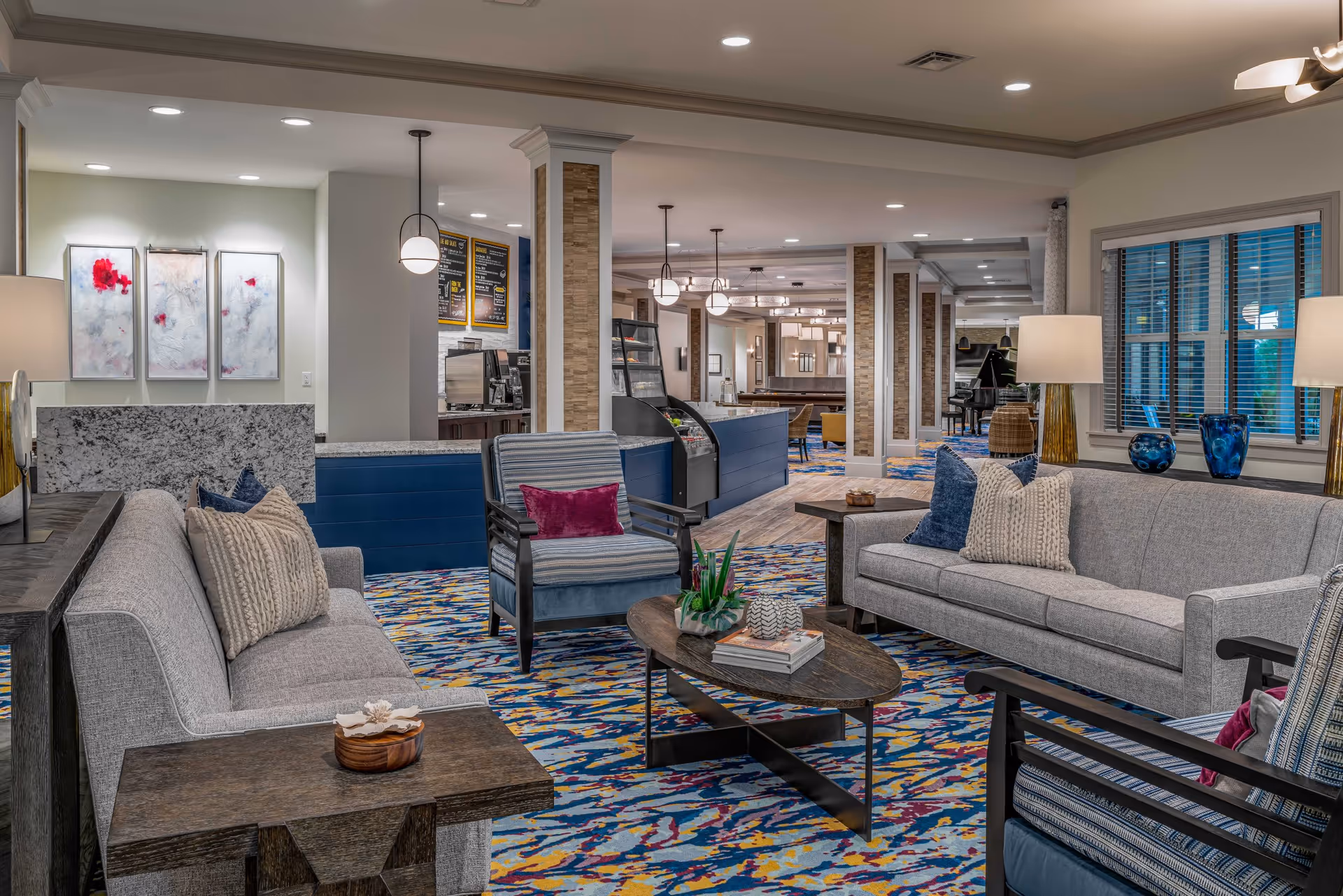 A spacious and well-lit living room area in Discovery Village Trinity featuring two gray sofas with decorative pillows, two striped armchairs with purple cushions, a wooden coffee table with books and a plant, side tables with lamps, colorful patterned carpet, and large windows with blinds. In the background, there is a counter area with a display case and menu boards, and further back, a piano and additional seating are visible.