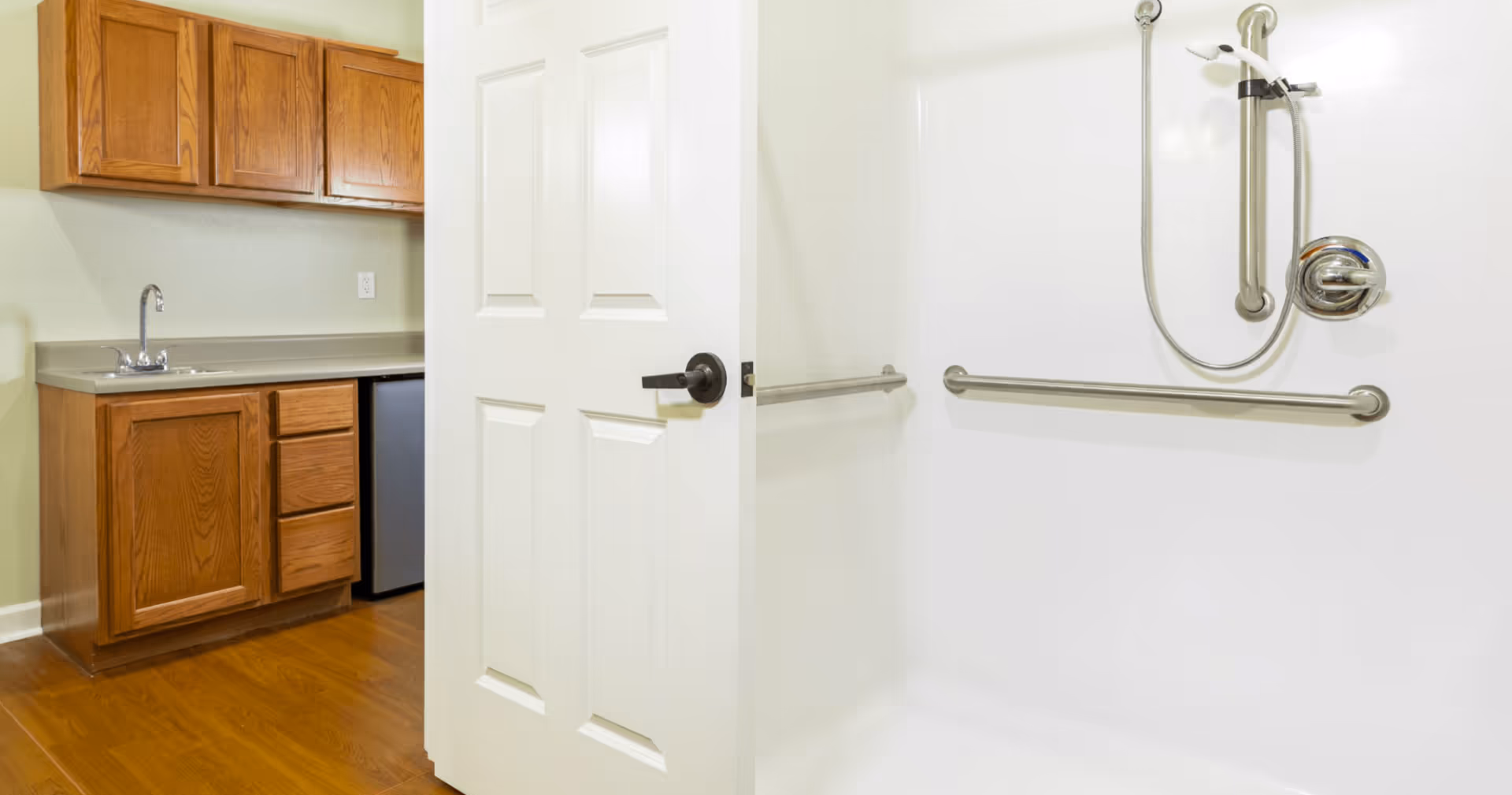 View of a bathroom with a white door partially open revealing a shower area with stainless steel grab bars and a handheld showerhead. Adjacent to the bathroom is a kitchenette area with wooden cabinets, a countertop, a sink, and a small refrigerator.