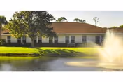 Single-story residential building with a brown roof and row of windows facing a pond with a fountain and grassy lawn.