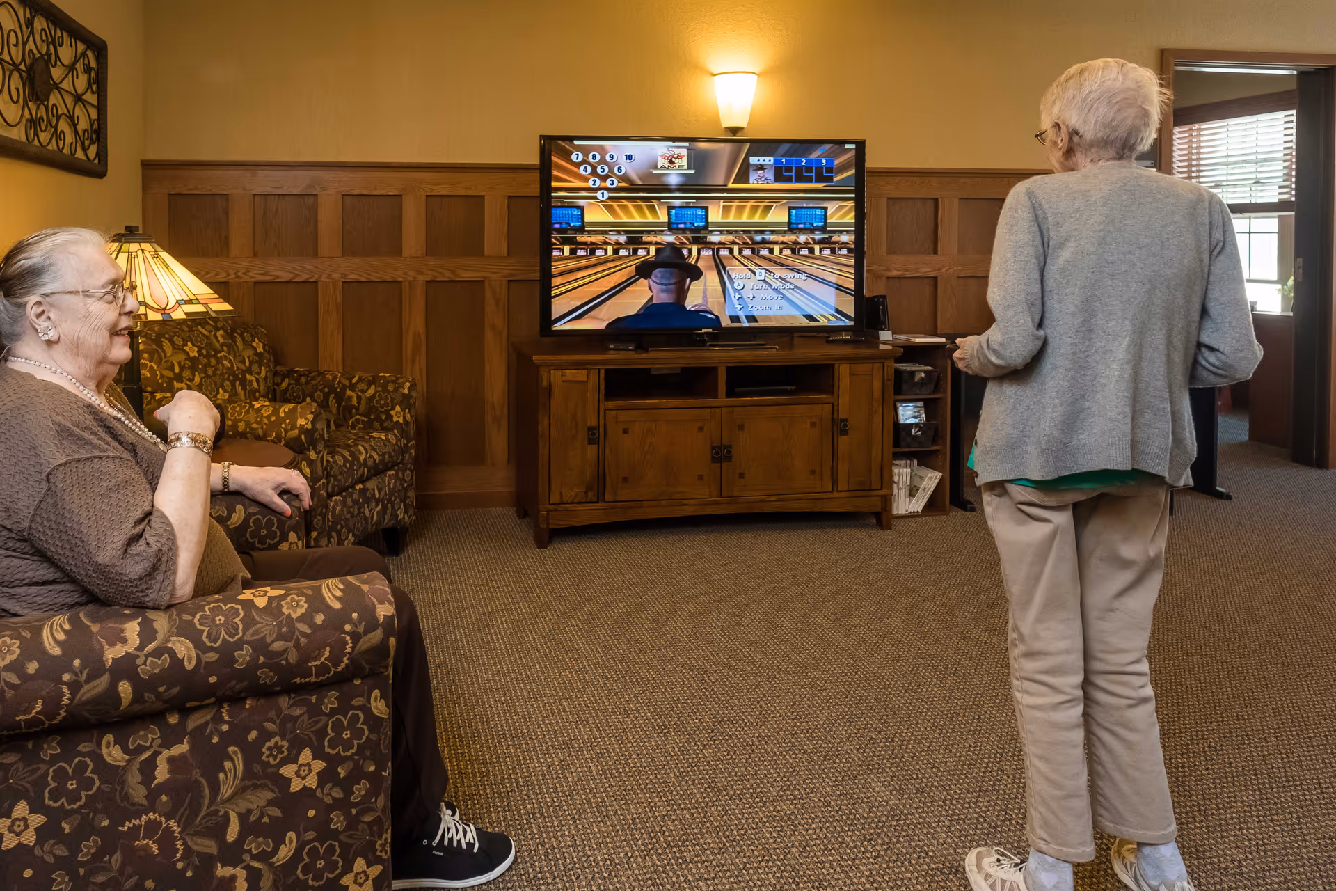 Two elderly women in a cozy living room setting; one woman is sitting on a floral-patterned armchair smiling, while the other woman stands facing a television screen playing a bowling video game. The room has wood paneling on the walls, a carpeted floor, and a wooden TV stand.