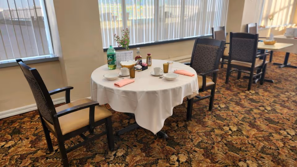 A dining area with round tables covered in white tablecloths, set with cups, bowls, napkins, and condiments. The room has large windows with vertical blinds and patterned carpet flooring. Several wooden chairs with cushioned seats surround the tables.