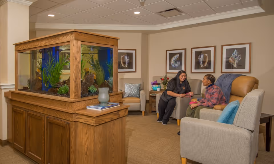 A cozy seating area in a senior living facility with a large wooden-framed aquarium filled with plants and fish. Two women are sitting and engaging in conversation on comfortable chairs. The walls are decorated with framed pictures of seashells.