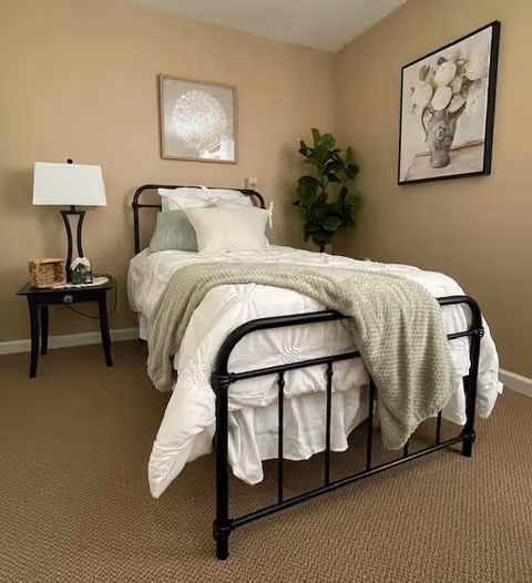 A neatly made single bed with a black metal frame, white bedding, and a light green knitted throw blanket draped over it. Next to the bed is a black nightstand with a white lamp, a small basket, and a decorative item. The room has beige walls with two framed floral artworks and a green potted plant in the corner.